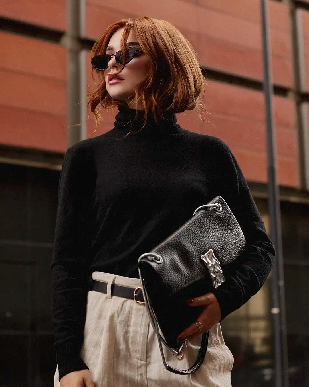 Mujer con look elegante, suéter negro de cuello alto, bolso negro con detalle metálico y tacones negros de Steve Madden, estilo elegante y sofisticado.
