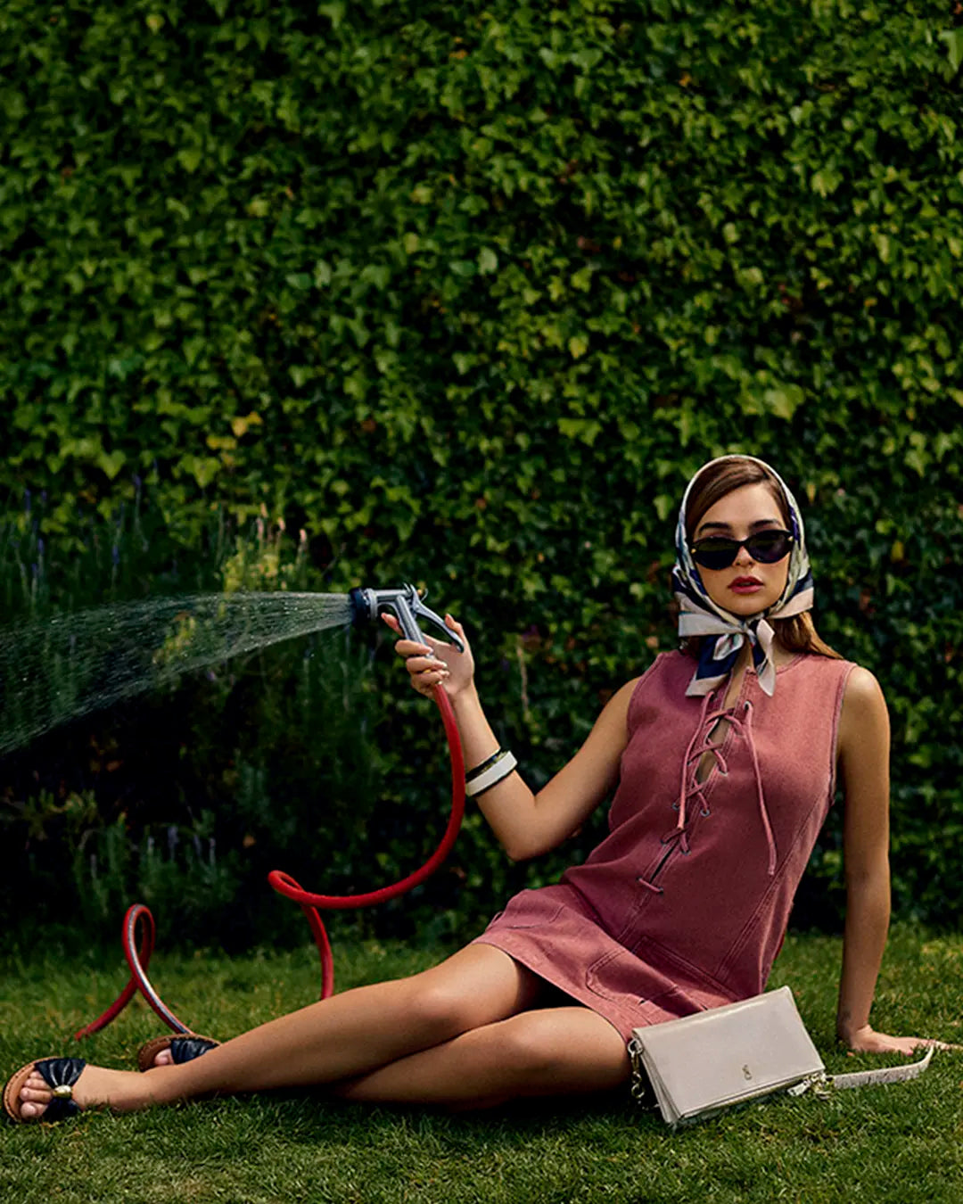 Mujer vistiendo vestido corto rosa sin mangas con detalle de cordones al frente, sandalias planas negras con esfera dorada al centro y gafas de sol Steve Madden, gafas de sol negras, con un look fresco elegante y sofisticado.